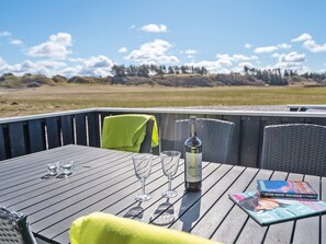 A wooden table with two wine glasses, a bottle of wine, and a magazine on a deck with a view of a grassy field and trees.