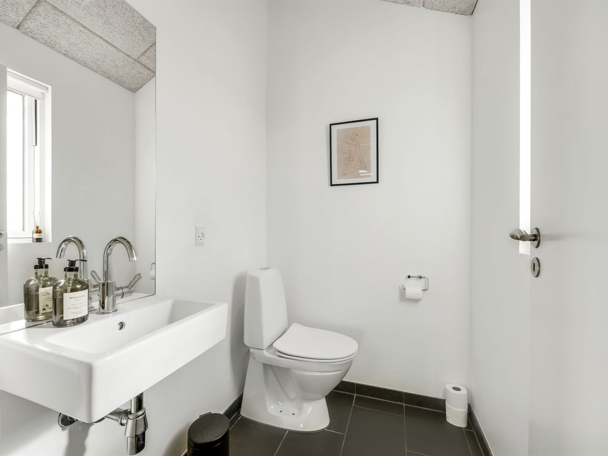 Ein modernes Badezimmer mit einem weißen Waschbecken, zwei Wasserhähnen, einer Toilette, einem gerahmten Bild an der Wand und einer Tür mit Schlüsselloch.