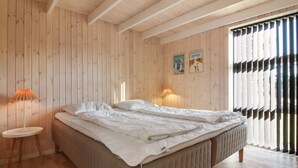 Een slaapkamer met een wand van houten latten, een bed met witte beddengoed, twee bedlampen en een raam met zwart-wit gestreepte jaloezieën.