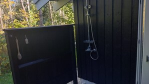 Een houten doucheomranding buiten, met een metalen tuinslang en douchekop, omgeven door kiezelstenen en een houten terras.