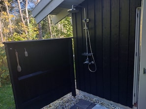 A wooden outdoor shower enclosure with a metal hose and showerhead, surrounded by pebbles and a wooden deck.