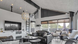 A modern living room with a sofa, coffee table, and chairs. There is a fireplace, a TV, and a large window.