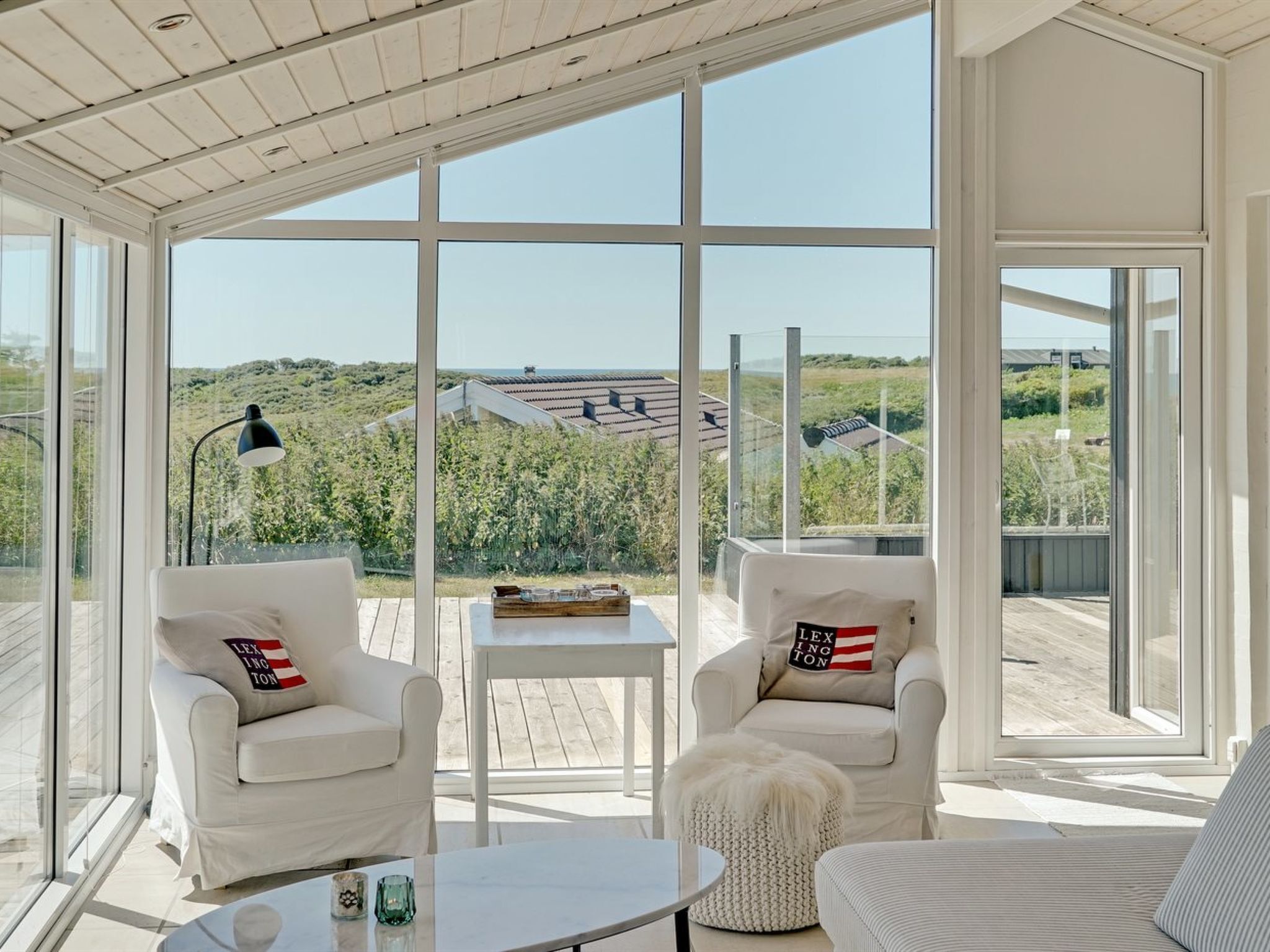 En rummelig balkon med hvide møbler og store vinduer, der giver udsigt over et landligt landskab.
