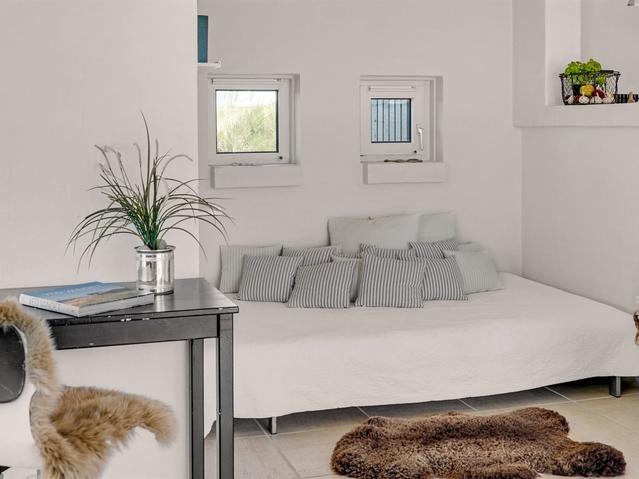 Ein modernes Schlafzimmer mit einem großen Bett, zwei Fenstern, einem kleinen Tisch mit Pflanze und einem Fellteppich.