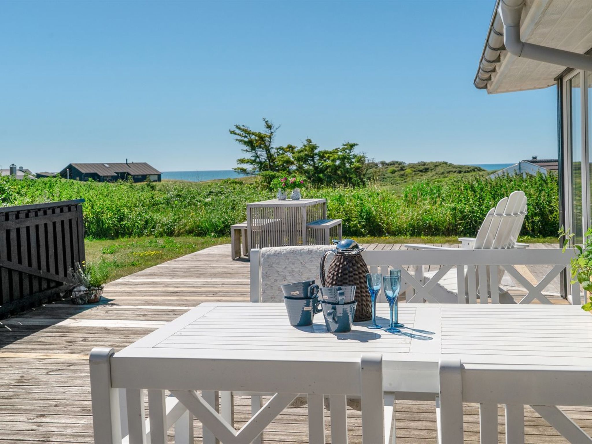Une terrasse en bois avec une table et des chaises, d’où l’on peut admirer un paysage côtier verdoyant, avec des maisons qui se dessinent à l’horizon.