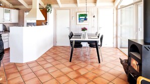 Amplia sala de estar con piso de baldosas, una mesa de comedor con sillas, una chimenea y una zona de cocina con gabinetes blancos y una planta colgante.