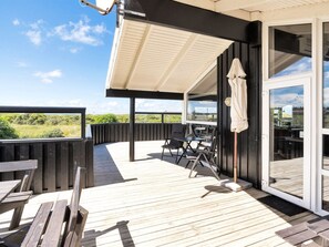 Une terrasse en bois avec des meubles d’extérieur, un espace salon couvert et une porte vitrée donnant sur une pièce.