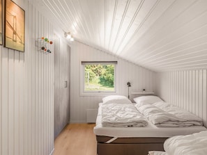 Ein Schlafzimmer mit geneigter Decke, einem Fenster mit Blick ins Grüne, einem Bett mit weißer Bettwäsche und einer Wandleuchte.