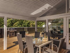 Une terrasse extérieure avec un patio fermé par des baies vitrées, des meubles en osier, un barbecue et une piscine visible à travers le verre.