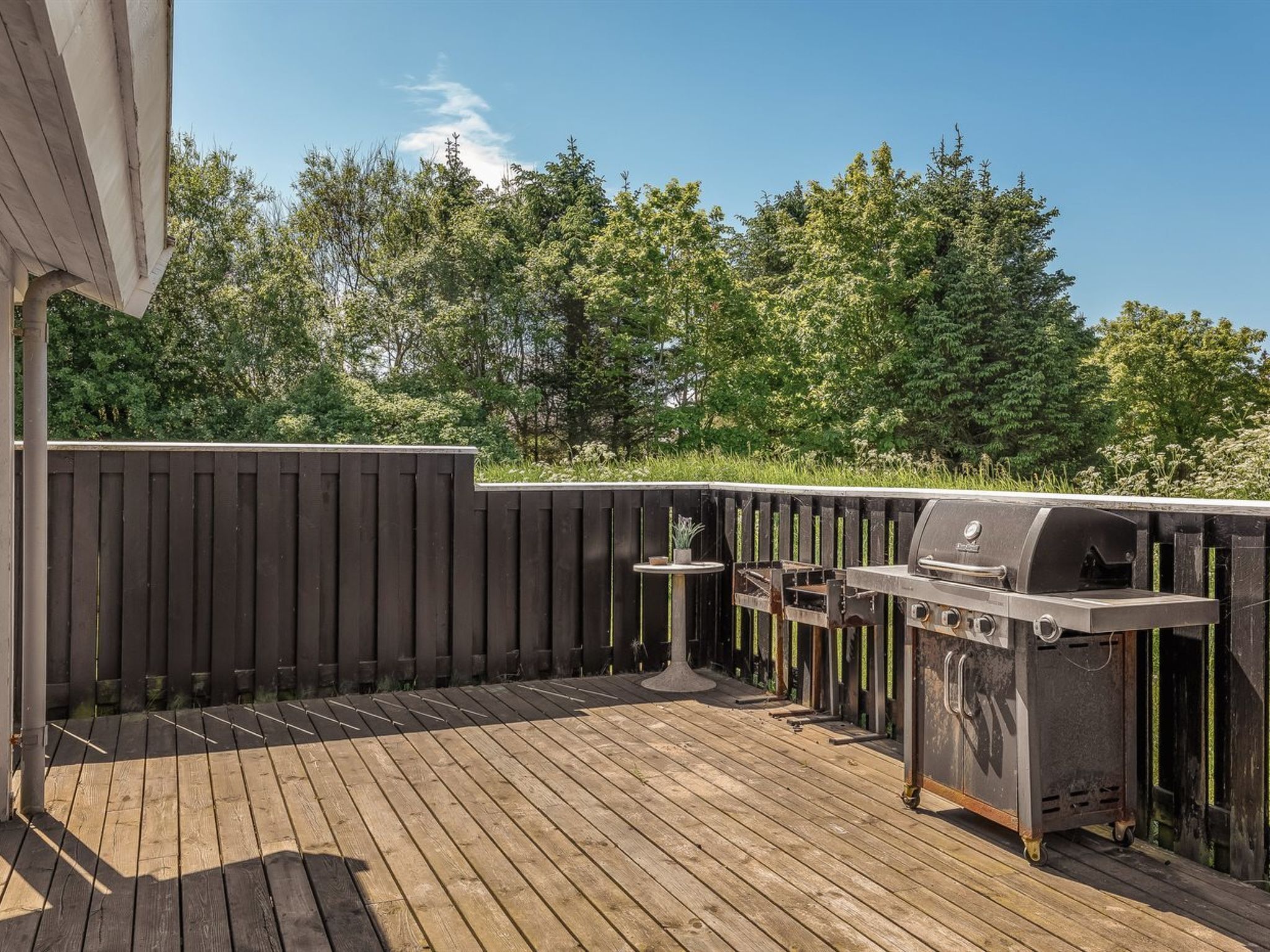 Una terraza de madera con una parrilla y una mesa, rodeada por una valla de madera y árboles.