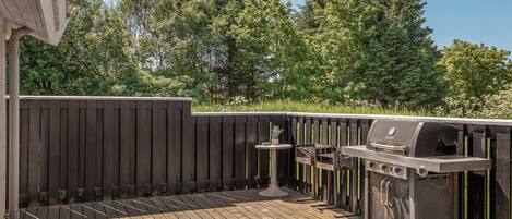 Una terraza de madera con una parrilla y una mesa, rodeada por una valla de madera y árboles.