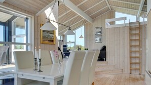 Une pièce spacieuse avec un plafond haut, des poutres apparentes, une table à manger avec des chaises et une vue dégagée sur l’extérieur grâce à de grandes fenêtres.