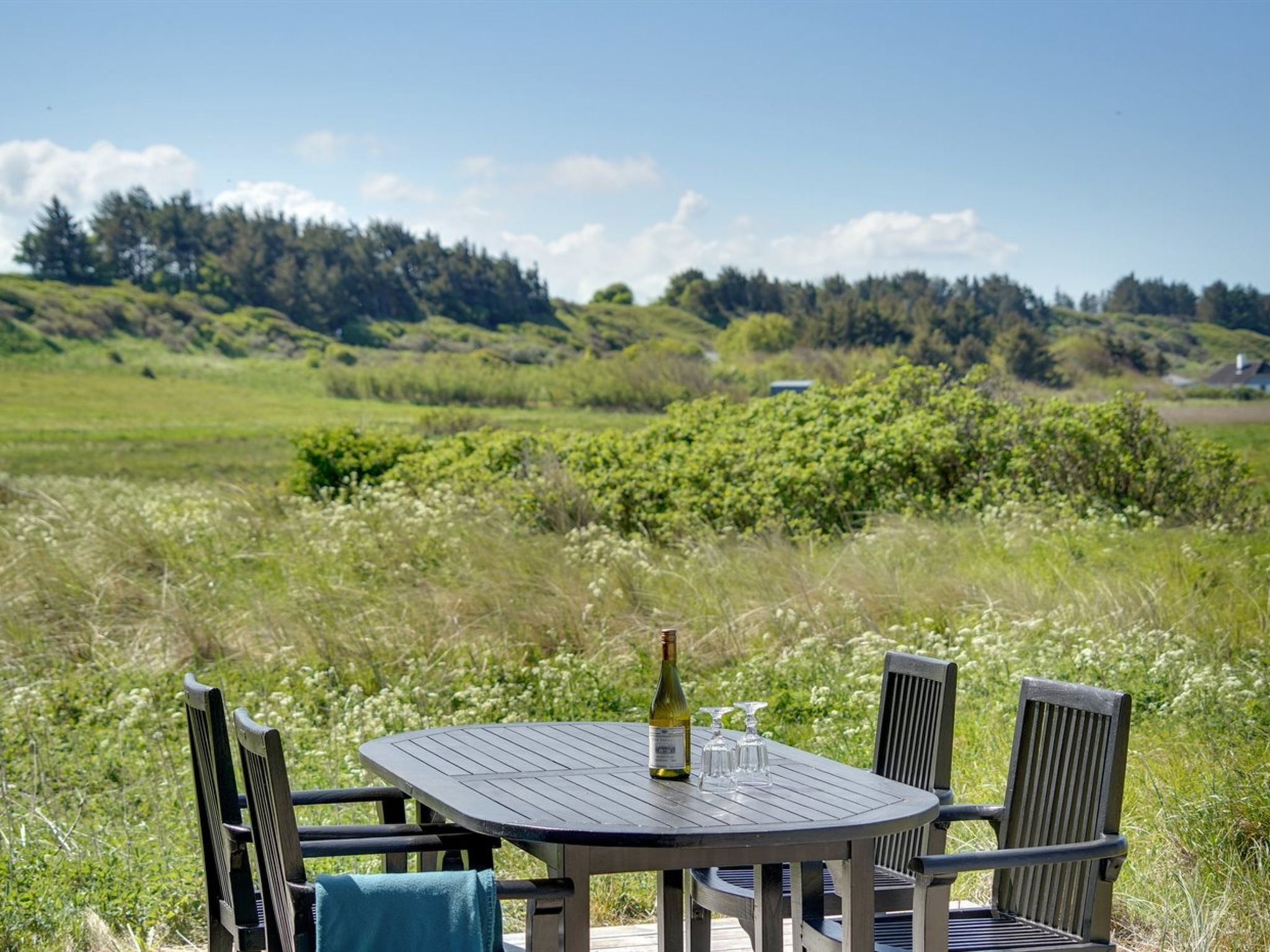 A round wooden table with four chairs, a bottle of wine, and two glasses on it, set outdoors with a grassy field and trees in the background.