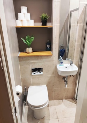 Compact bathroom with a toilet, sink, and shower area, featuring a shelf for essentials.