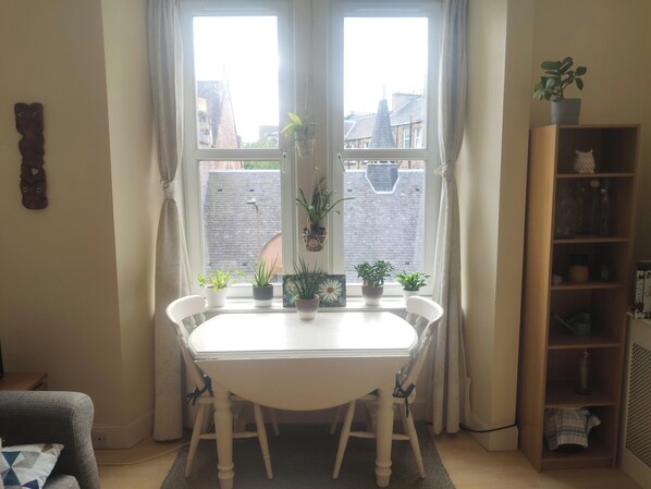 Enjoy a bright and airy room with a large window offering views of the surrounding buildings, a dining table, and a bookshelf.