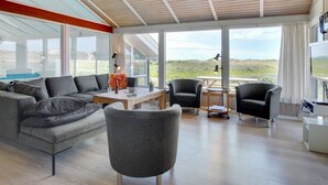 A spacious living area with a large sofa, two armchairs, a coffee table, and a TV. The room has large windows offering a view of the outdoors.