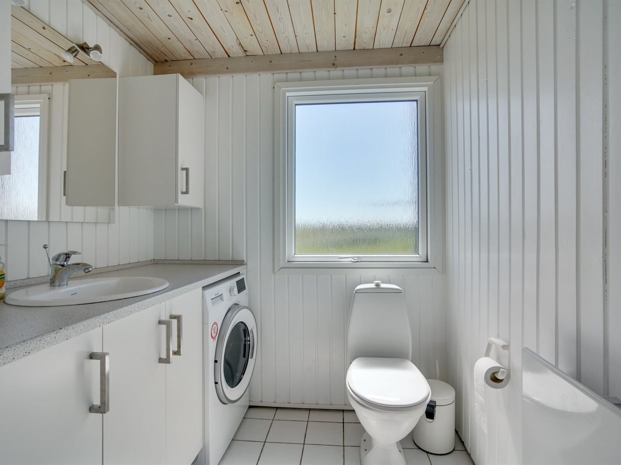 A compact, white-walled bathroom with a built-in washing machine, a toilet, and a window providing a view of the outdoors.