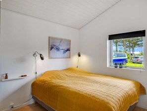 Une chambre à coucher avec un lit, deux lampes de chevet, un tableau, une fenêtre donnant sur des voitures garées et une étagère avec divers objets.