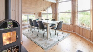 Une pièce chaleureuse avec un poêle à bois, une table à manger avec des chaises et de grandes fenêtres donnant sur l’extérieur.