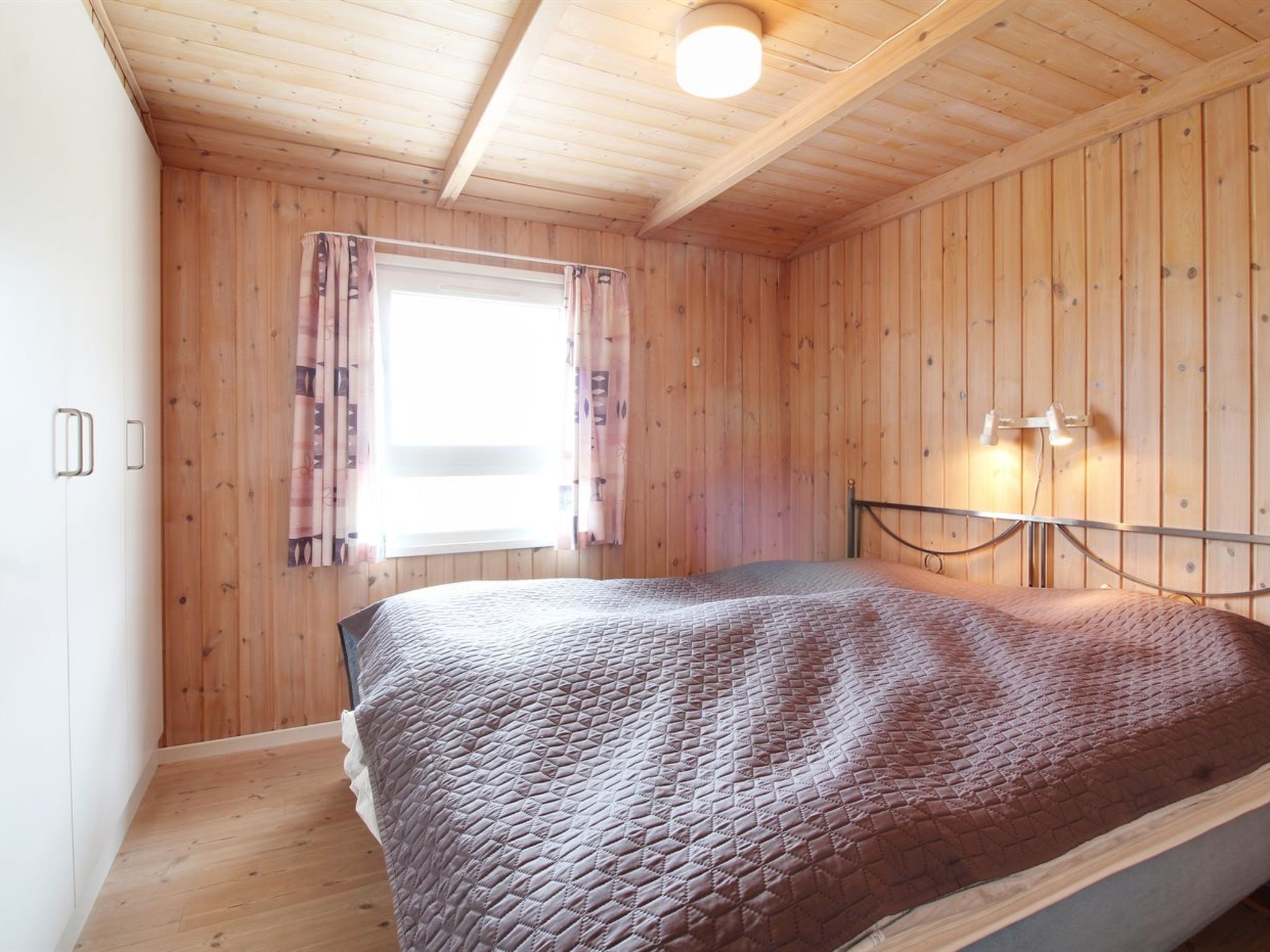 Et værelse med trævægge, en seng med quiltet dyne, et vindue med gardiner og en vægmonteret lampe.