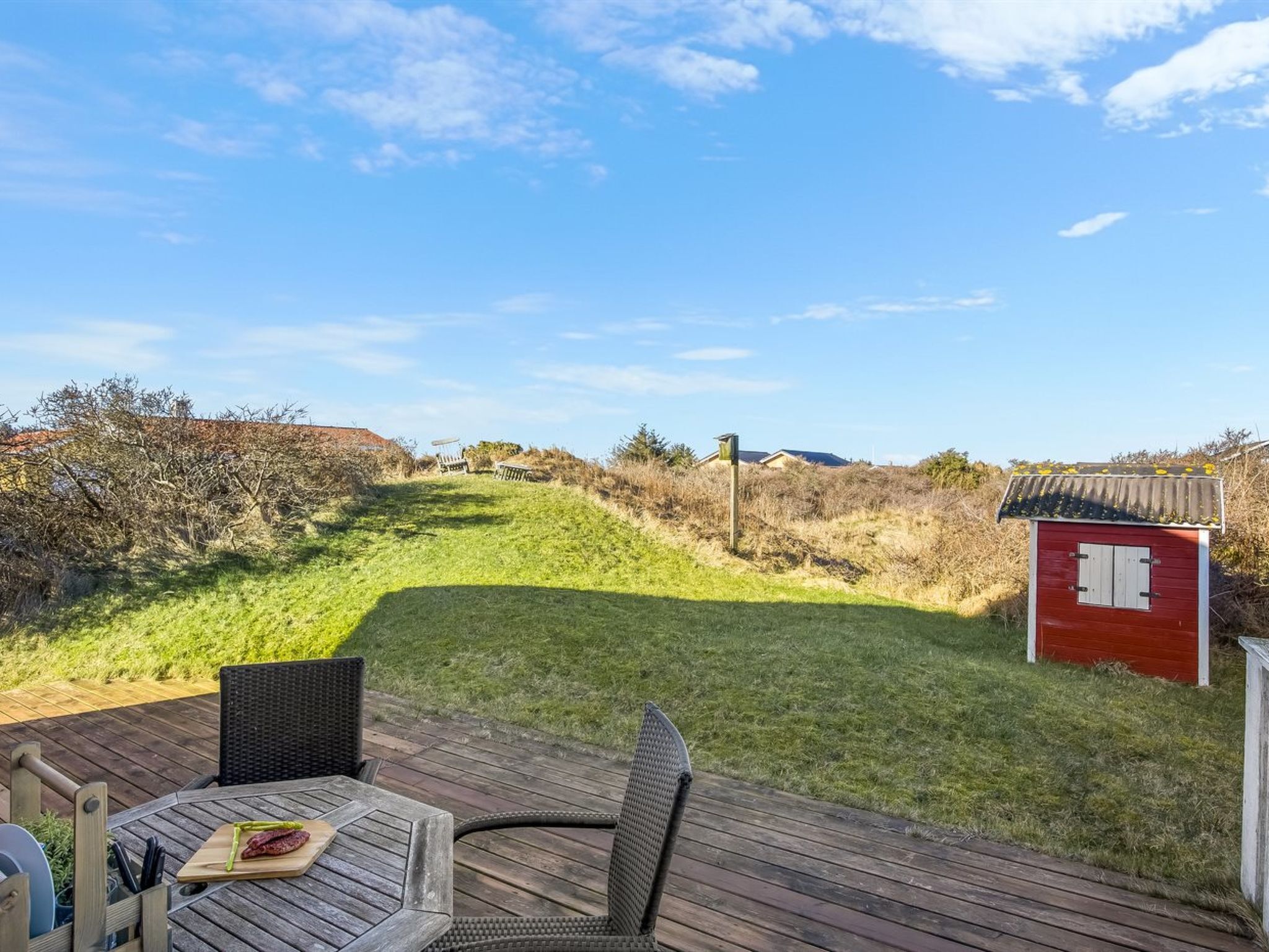 Une terrasse avec une table et des chaises, un petit abri de jardin rouge et, au fond, une pente herbeuse.