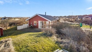 Une maison rouge avec une terrasse, entourée d’herbe sèche et de buissons.