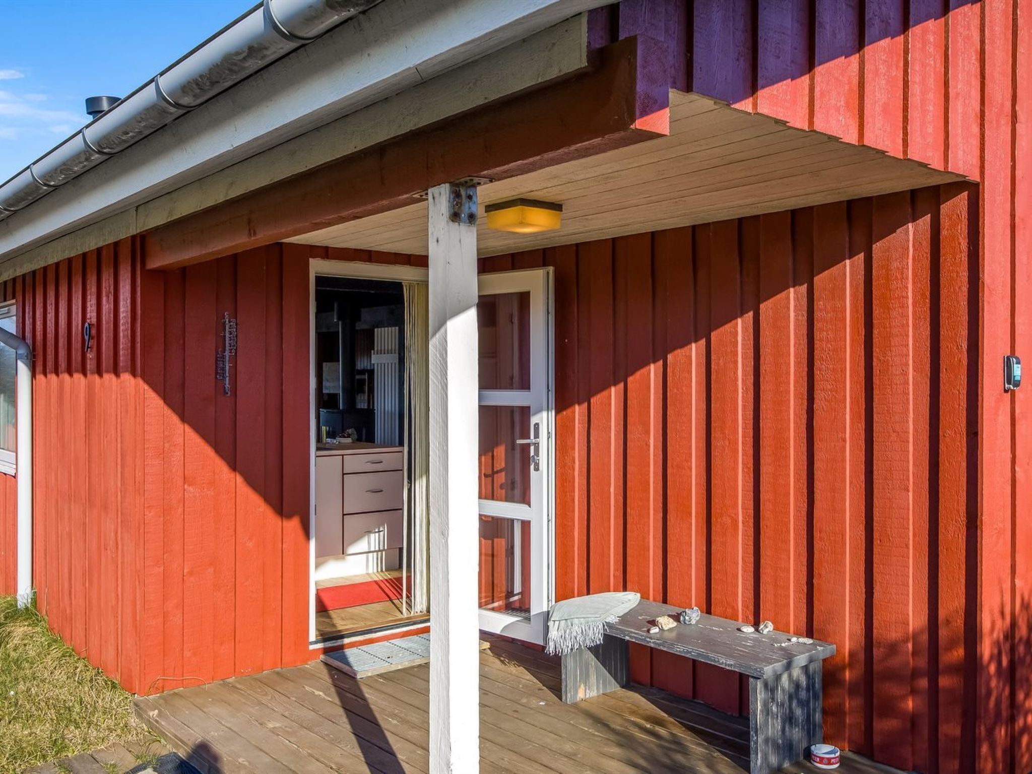 Un bâtiment rouge avec une terrasse en bois, une porte blanche et une petite table ornée de pierres.
