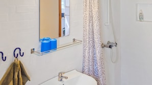 Ein Badezimmer mit Waschbecken, Spiegel und Dusche. Blaue Seifenspender sind an der Wand angebracht, und ein Handtuch hängt an einem Haken.