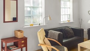 Ein Wohnzimmer mit einer schwarzen Ledersofa, einem Holzstuhl, einem kleinen Holztisch, einem roten Beistelltisch mit Korb und zwei Fenstern mit Jalousien.