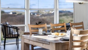 A dining area with a large table, wooden chairs, and a view of the outdoors through large windows.