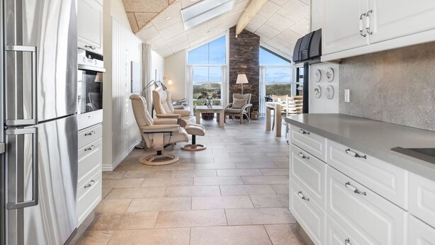 A modern kitchen with white cabinetry, a stone countertop, and a built-in oven. There are two beige chairs and a table with a lamp.