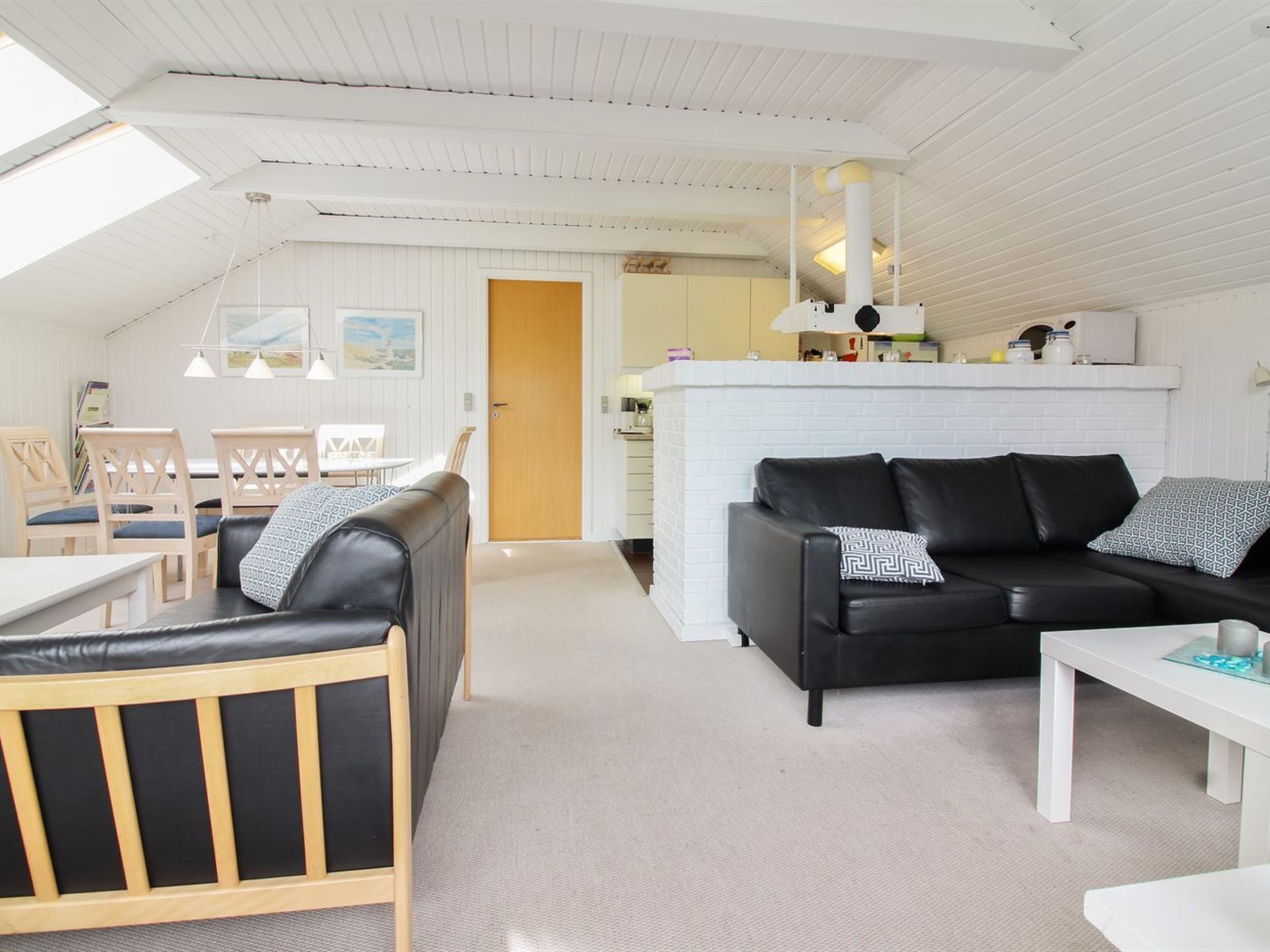 Un salon moderne avec un canapé en cuir noir, des chaises en bois, une table basse blanche et un coin cuisine doté d’un mur de briques blanches.