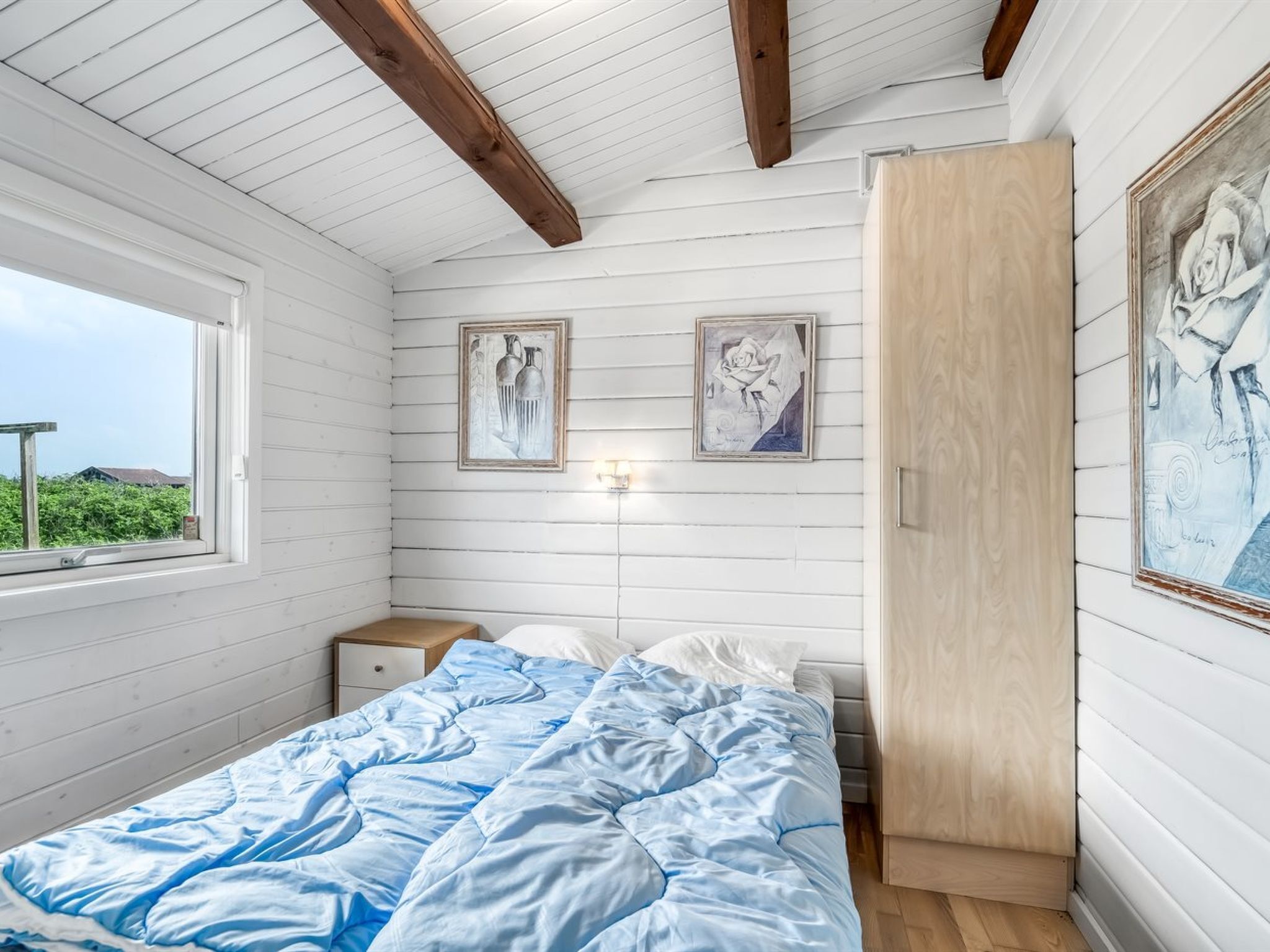 Ein Schlafzimmer mit Holzboden, einem Bett mit blauer Steudecke, zwei gerahmten Kunstwerken an der Wand und einem Holzschrank.