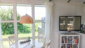 Un coin repas avec une table ronde, des chaises blanches et une lampe suspendue. On peut voir une bibliothèque remplie de livres et un téléviseur posé sur un meuble.