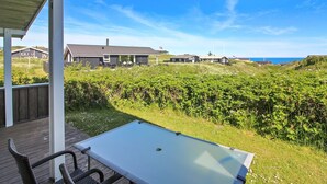 Une terrasse couverte, dotée d’une table et de chaises, donnant sur un espace verdoyant et offrant une vue sur les maisons et la mer.