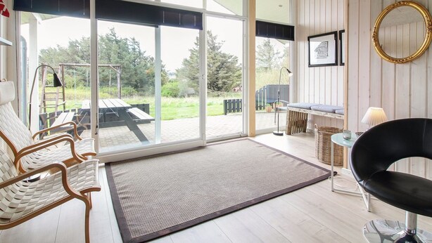 A spacious room with a large sliding glass door leading to an outdoor area, a wooden bench, a black chair, and a small table with a lamp. - "Herbiorn" - 403m from the sea by Interhome (Hjørring)