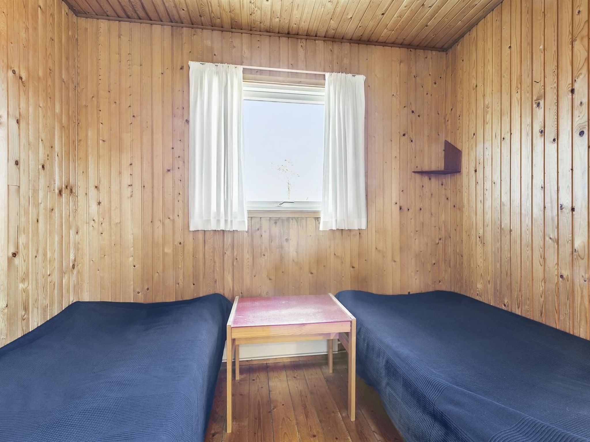 A room with wooden walls, two single beds, a small wooden table, and a window with white curtains.