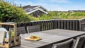 Eine Holzterrasse mit Tisch und Stühlen, ein Grill mit Essen und im Hintergrund Häuser.