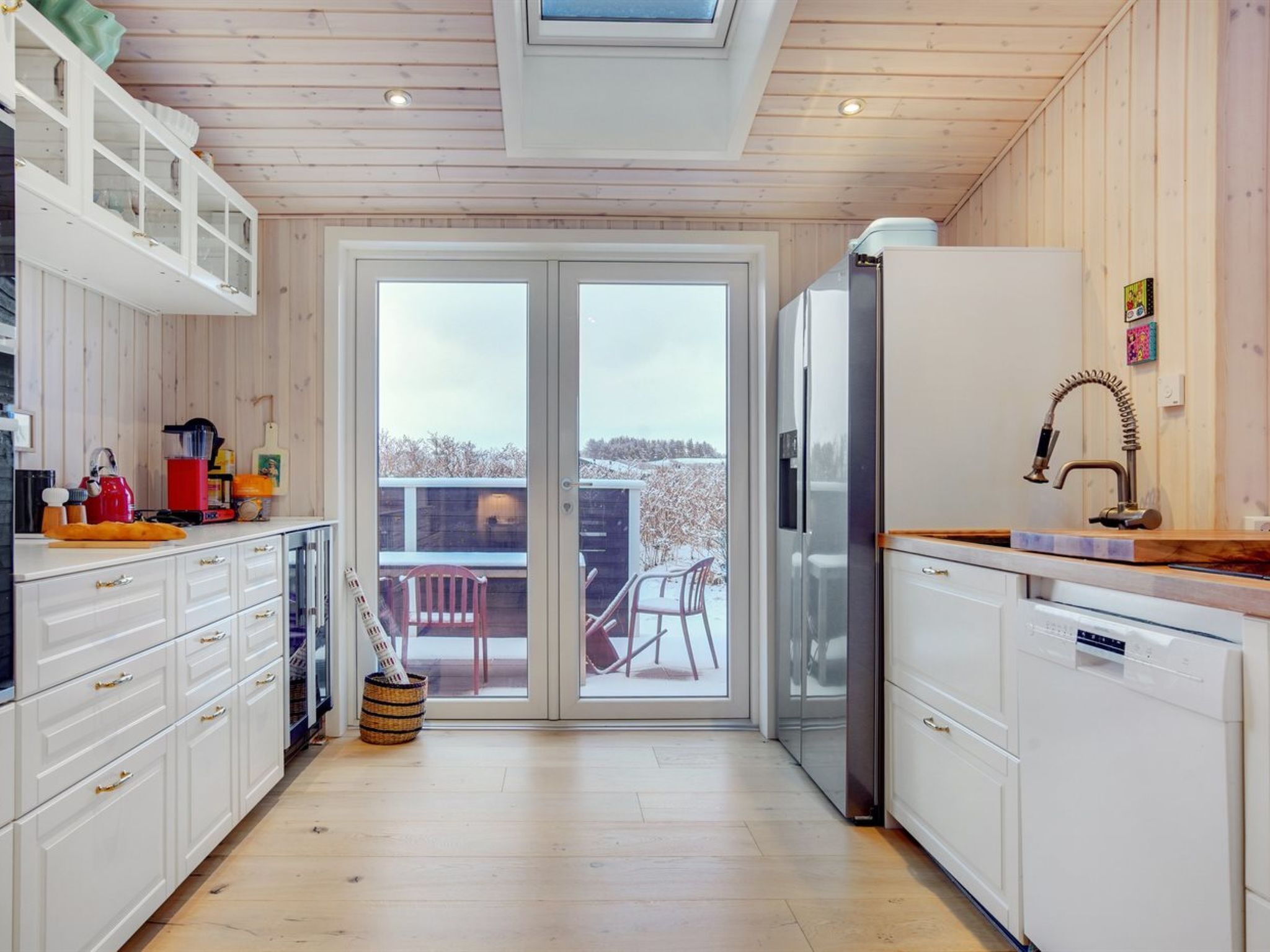 A modern kitchen with white cabinets, a stainless steel refrigerator, and a large window overlooking a snowy landscape.