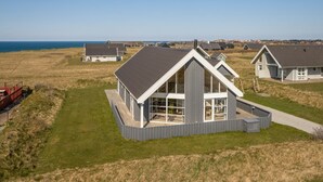 A modern coastal house with a large balcony, surrounded by a grassy field and a view of the sea.