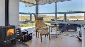 Een knus vertrek met een houtkachel, grote ramen, een comfortabele fauteuil en een kleine tafel.