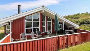 Une maison en bois rouge avec une grande fenêtre en verre, une cheminée et une terrasse dotée de chaises et d’une table.