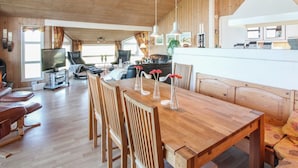 Un vaste espace de vie doté d’un plafond en bois, d’une table à manger avec des chaises, d’un canapé, d’un téléviseur et d’un coin cuisine avec un plan de travail blanc.