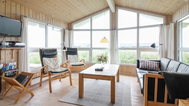 Une pièce spacieuse avec un plancher en bois, un canapé, deux fauteuils, une table et un téléviseur. De grandes fenêtres laissent entrer la lumière naturelle et offrent une vue sur la verdure environnante.