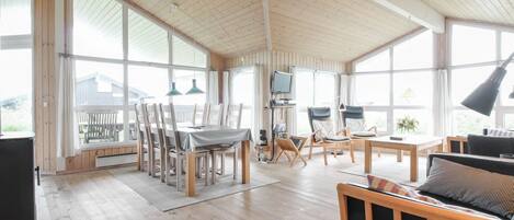 A spacious room with wooden floors, a dining area with chairs and a table, a sofa, and large windows providing natural light.