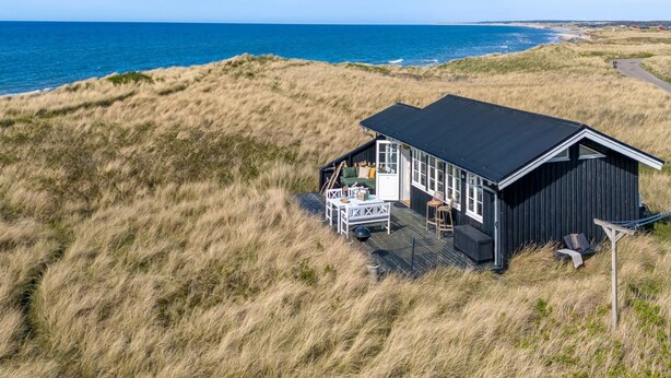 Una casa costera con terraza y zona de estar al aire libre, rodeada de dunas cubiertas de hierba y el océano.