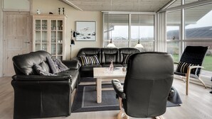 Una sala de estar moderna con un sofá de cuero negro, una mesa de centro de madera y un sillón negro. Hay un aparador con cristalería y un cuadro en la pared.