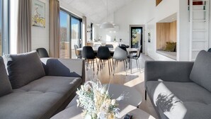 Een moderne woonkamer met grijze banken, een salontafel met een bloemstuk, en een eetgedeelte met een keuken op de achtergrond.