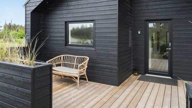 Een modern zwart houten gebouw met een schuin dak, een glazen deuropening en een houten terras met een rieten stoel.