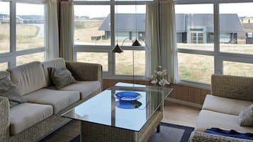 Un salon moderne avec une table basse en verre, des meubles en osier et de grandes fenêtres donnant sur la campagne.